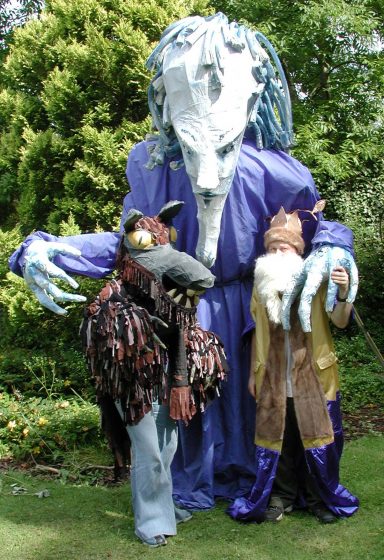 A giant blue puppet of a person stands behind a King and a wolf