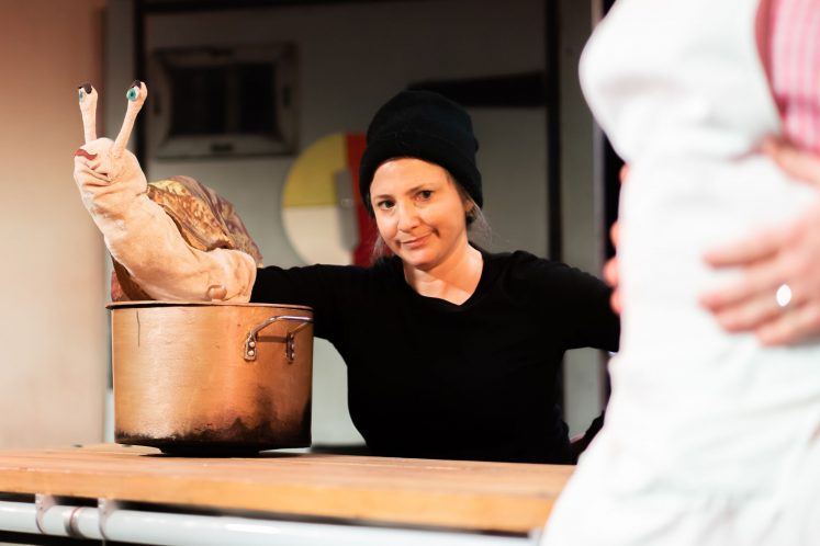 Puppeteer operates a snail puppet balanced on a saucepan. She is dressed in black and looks to the camera