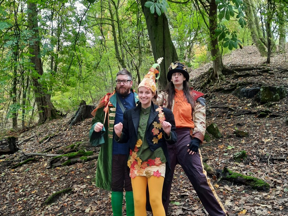 Three characters dressed in hats and clothing with a woodland theme stand together and smile at the camera