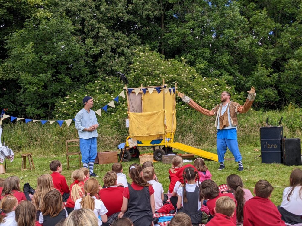 Two male actors perform outside to school children. They are dressed in blue and there is a yellow wooden cart parked infron of leafy green trees