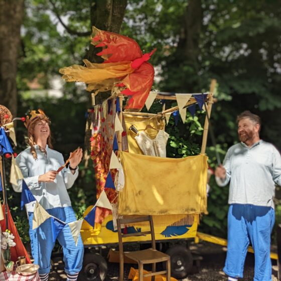 Two actors dressed in tones of blue against a wooden yellow cart. The one on the right operates a firebird puppet