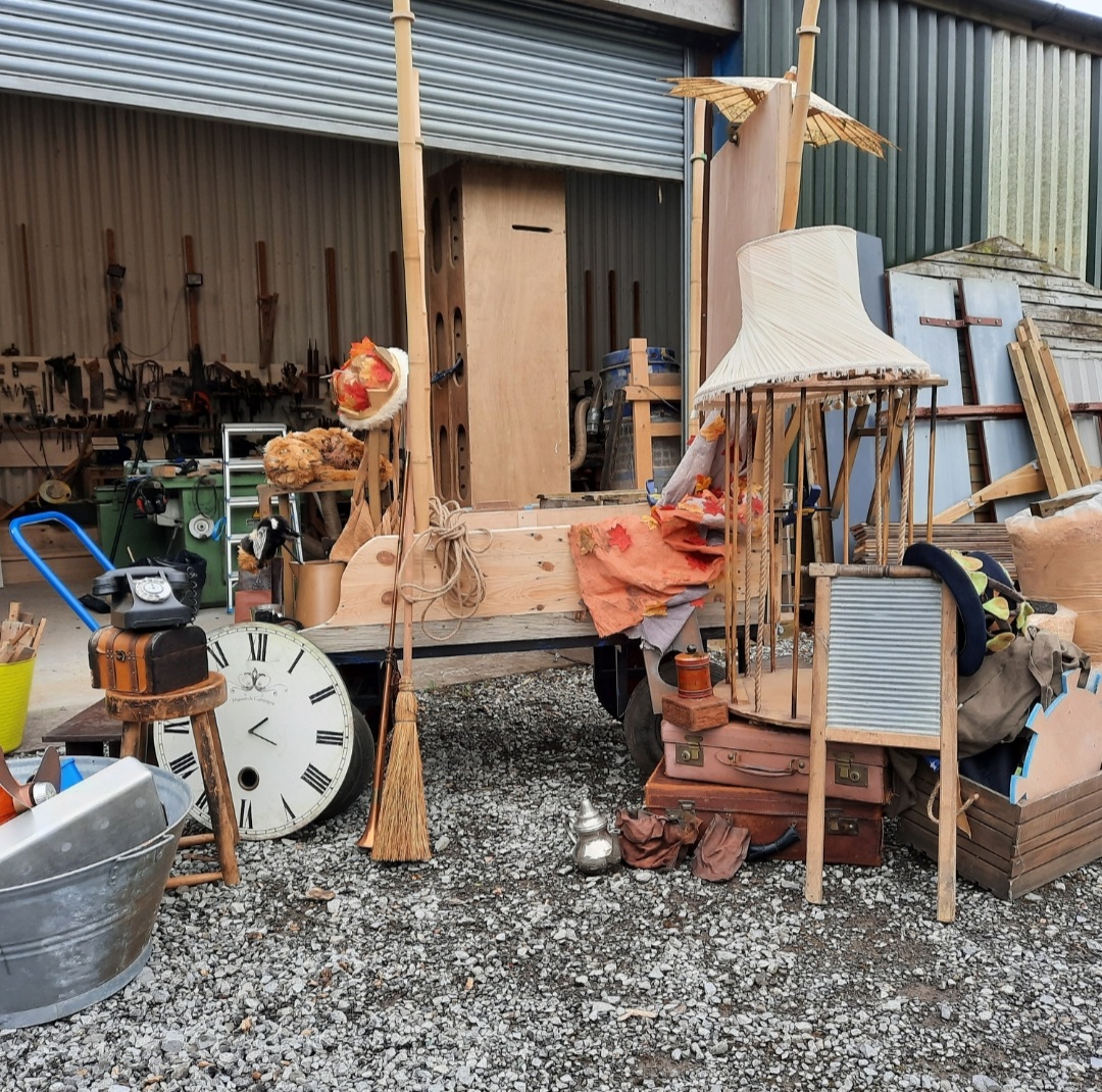 A cart with a jumble of objects all piled up around it. Old suitcases, lampshades, kitchenware make up the arranged items