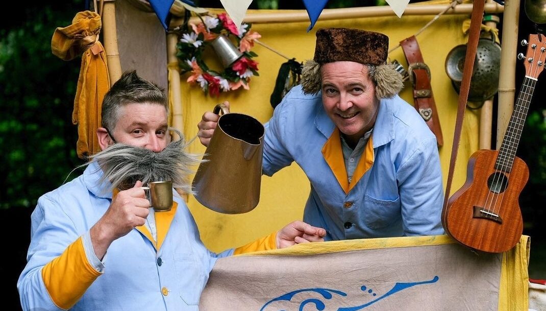 Two performers dressed in blue stand beside a bright yellow wagon filled with strange objects