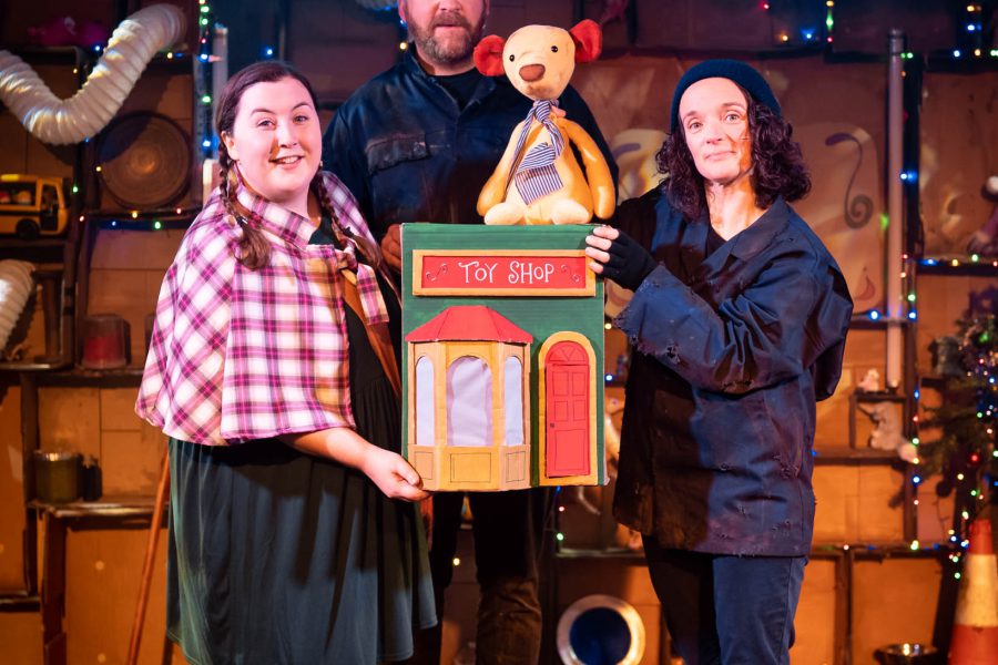 3 performers look out to the camera. They are on stage and are holding a toyshop with a teddy bear sitting on top.