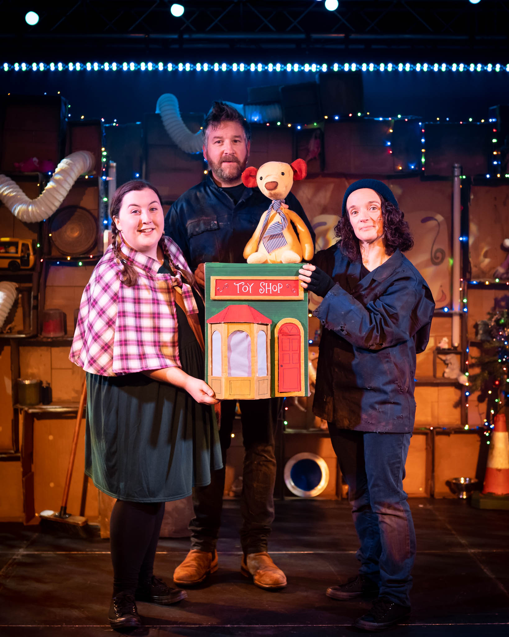 3 performers look out to the camera. They are on stage and are holding a toyshop with a teddy bear sitting on top.