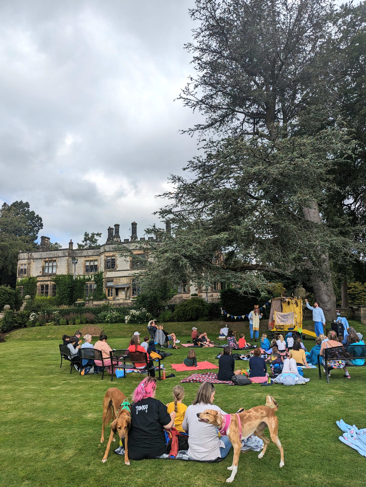 A theatrical performance is being held on the lawns of a grand house. The audience look on at the performance.