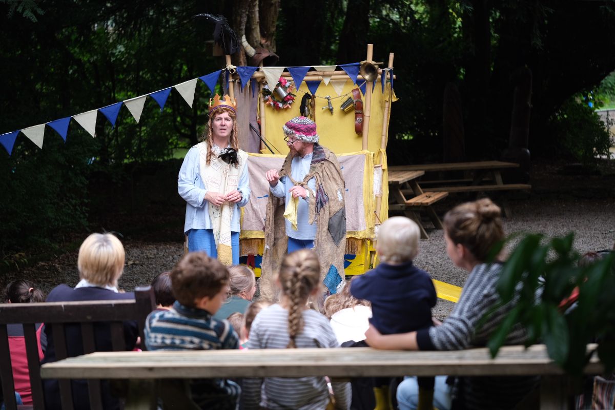 two actors tell a story to an audience. The image is taken from behind the audience and you can see the back of everyone's heads.