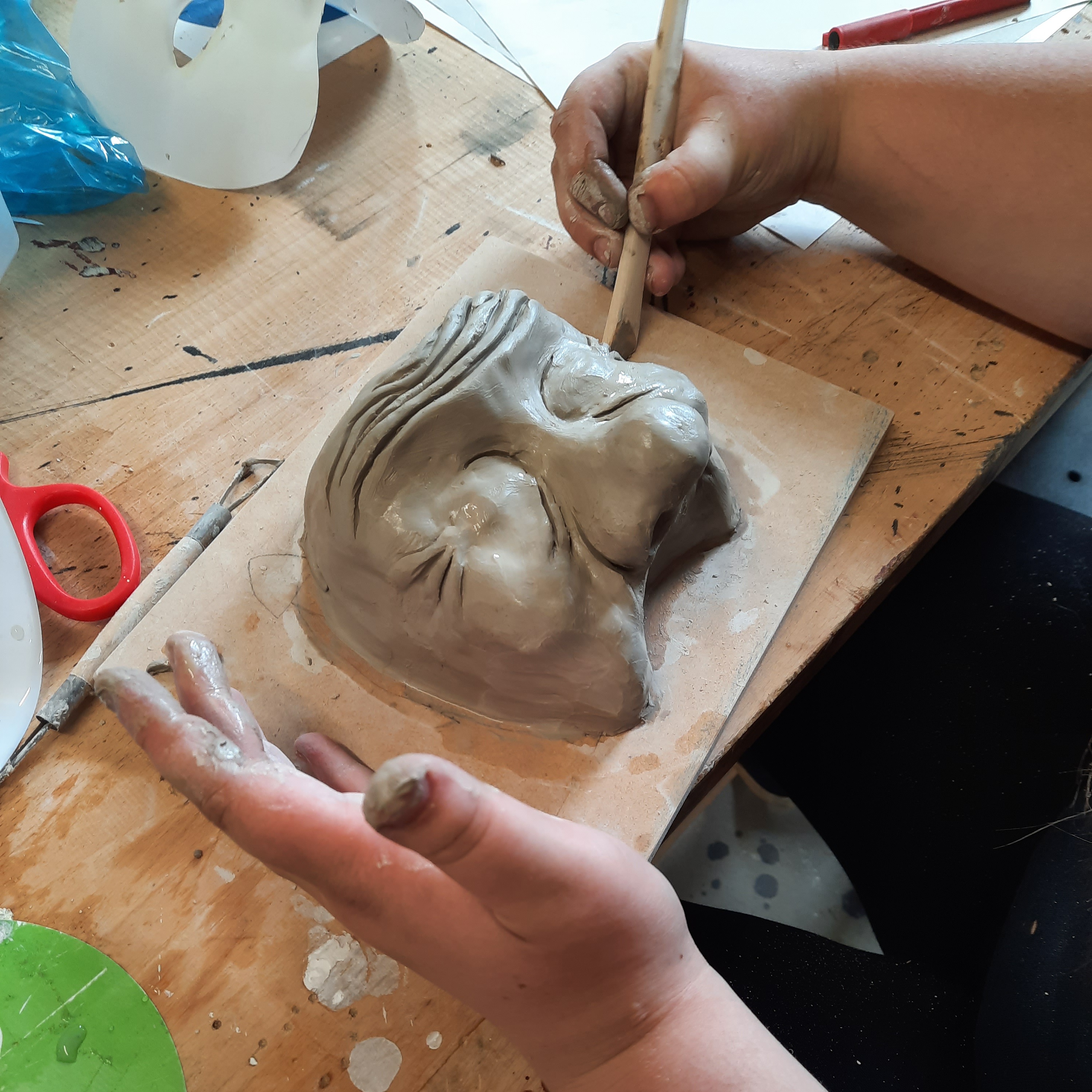 A close up of a student sculpting a clay face which has over the top features of a large nose and wrinkly forehead