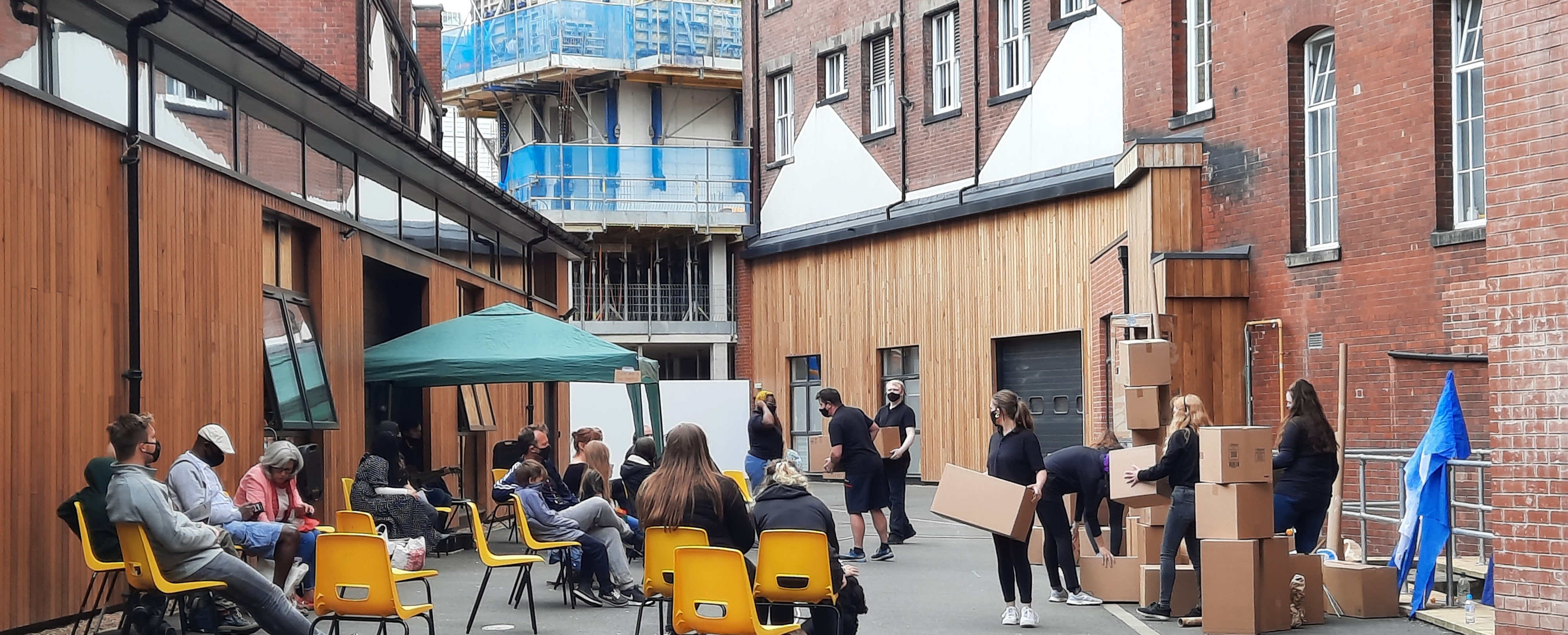 In a high walled courtyard, performers dressed in black carry boxes as an audience watch on