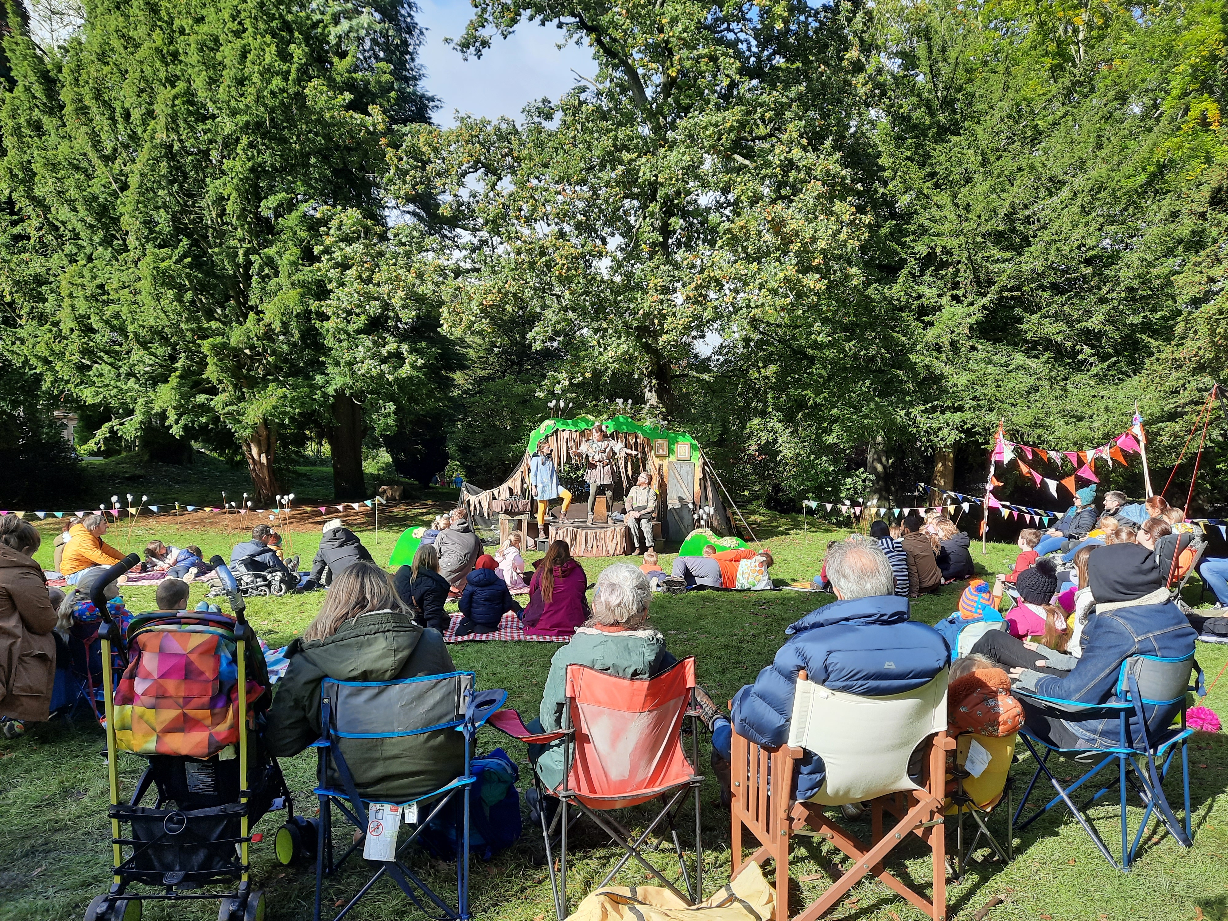 The photo is taken behind the audience. There is bunting a stage and a backdrop of greenleafed trees