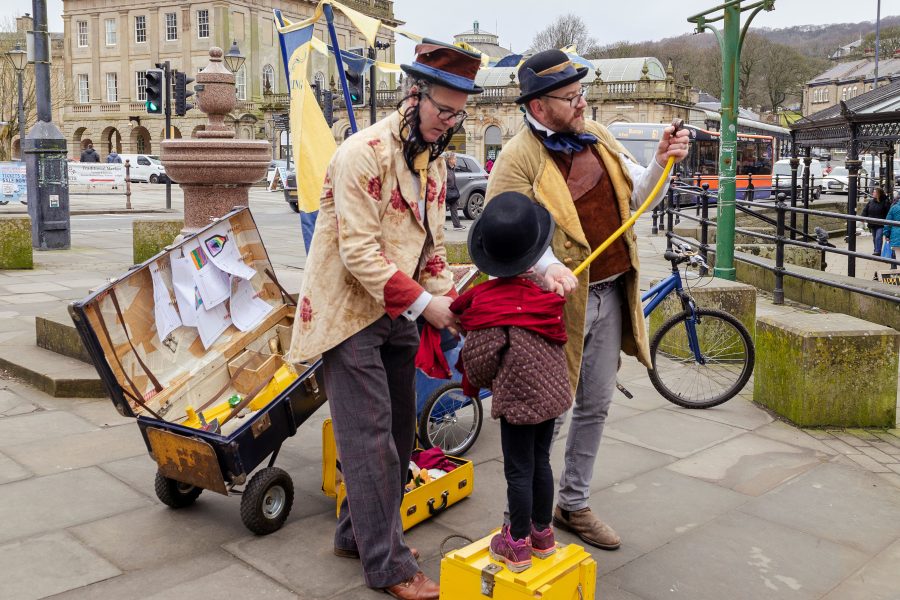 Two characters dressed as victoriana chemists are pretending to cure a small child who is stood on a yellow wooden box
