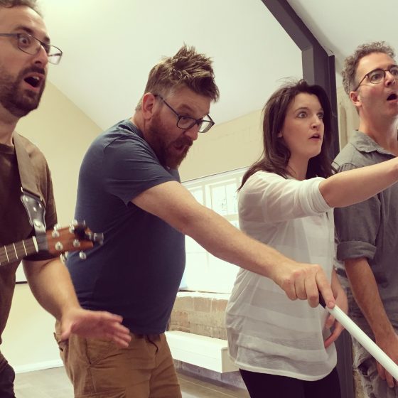 Four actors in a rehearsal room looking or pointing to their left with a shocked look on their faces