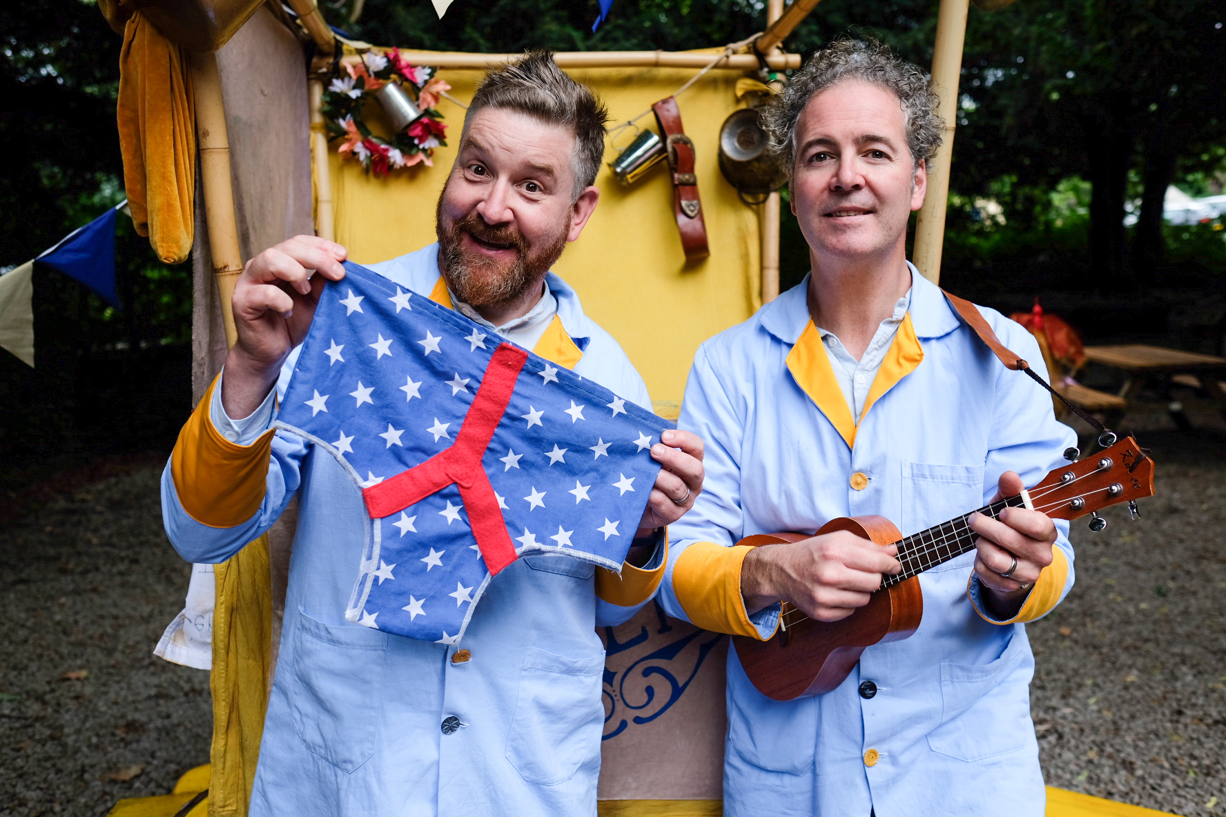 Two actors dressed in blue over coats stand in front of a yellow cart. One holds a ukelele and the other a big pair of blue starry pants