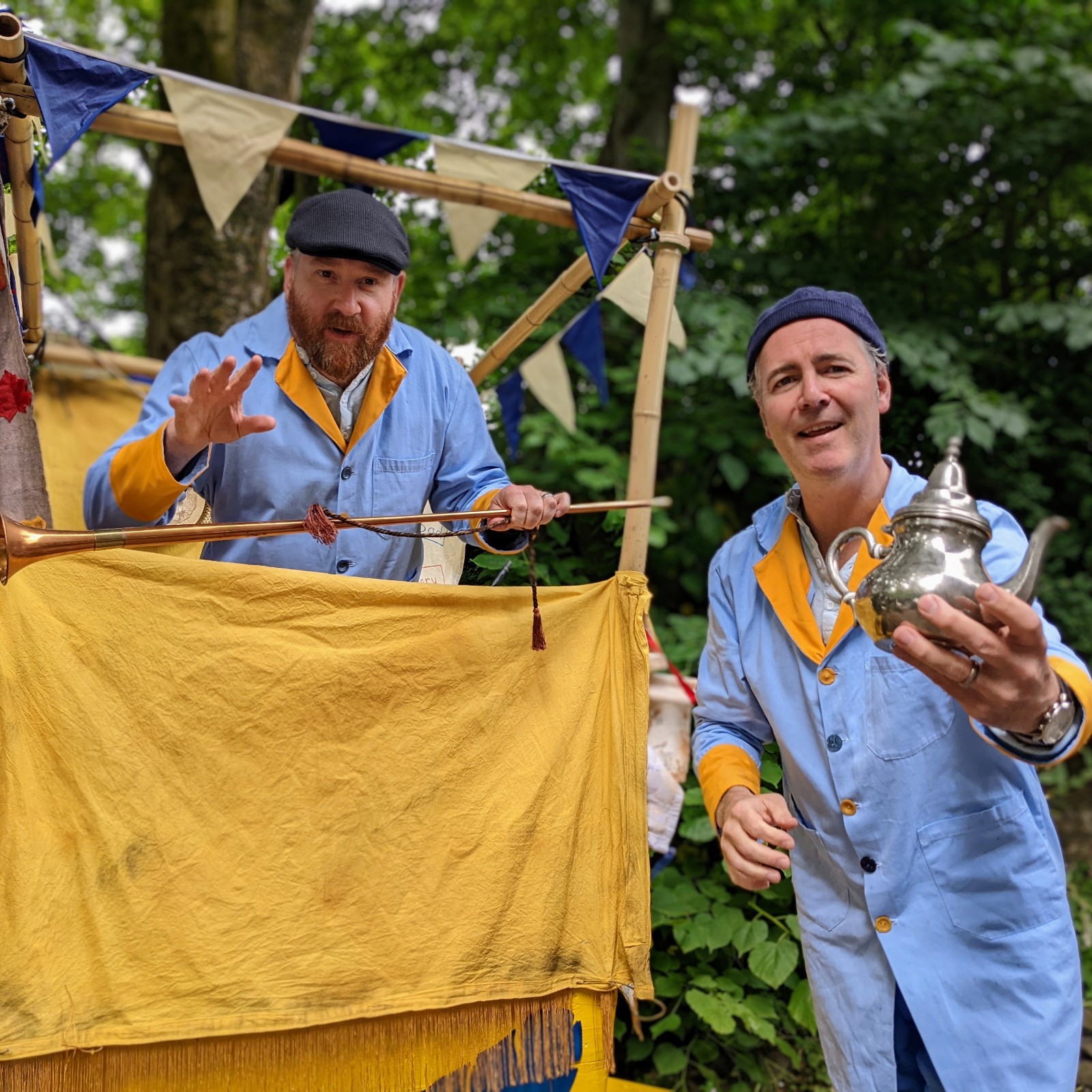 Two male performers dressed as sales assistants stand in front of a yellow cart. One holds a silver teapot towards the camera