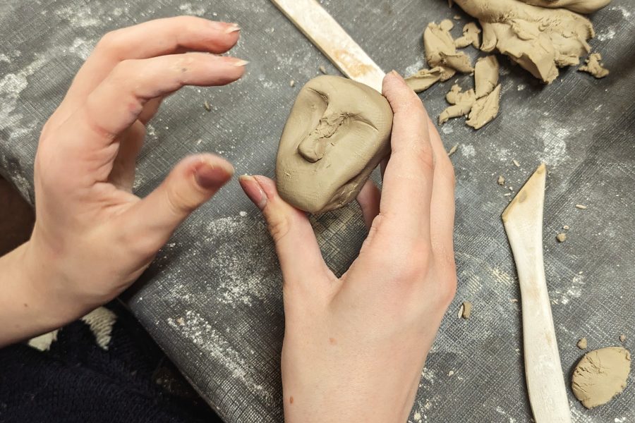 A persons hand sculpt a little clay face. There are clay tools and pieces of clay on the table surface below.