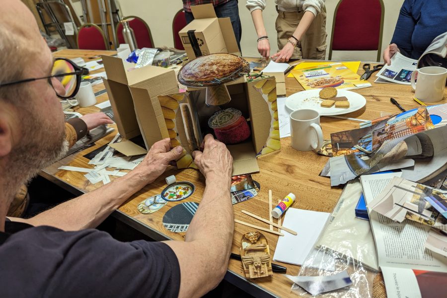 We see the shoulders of a man making a piece of artwork at a table. He is surrounded by magazine cuttings.