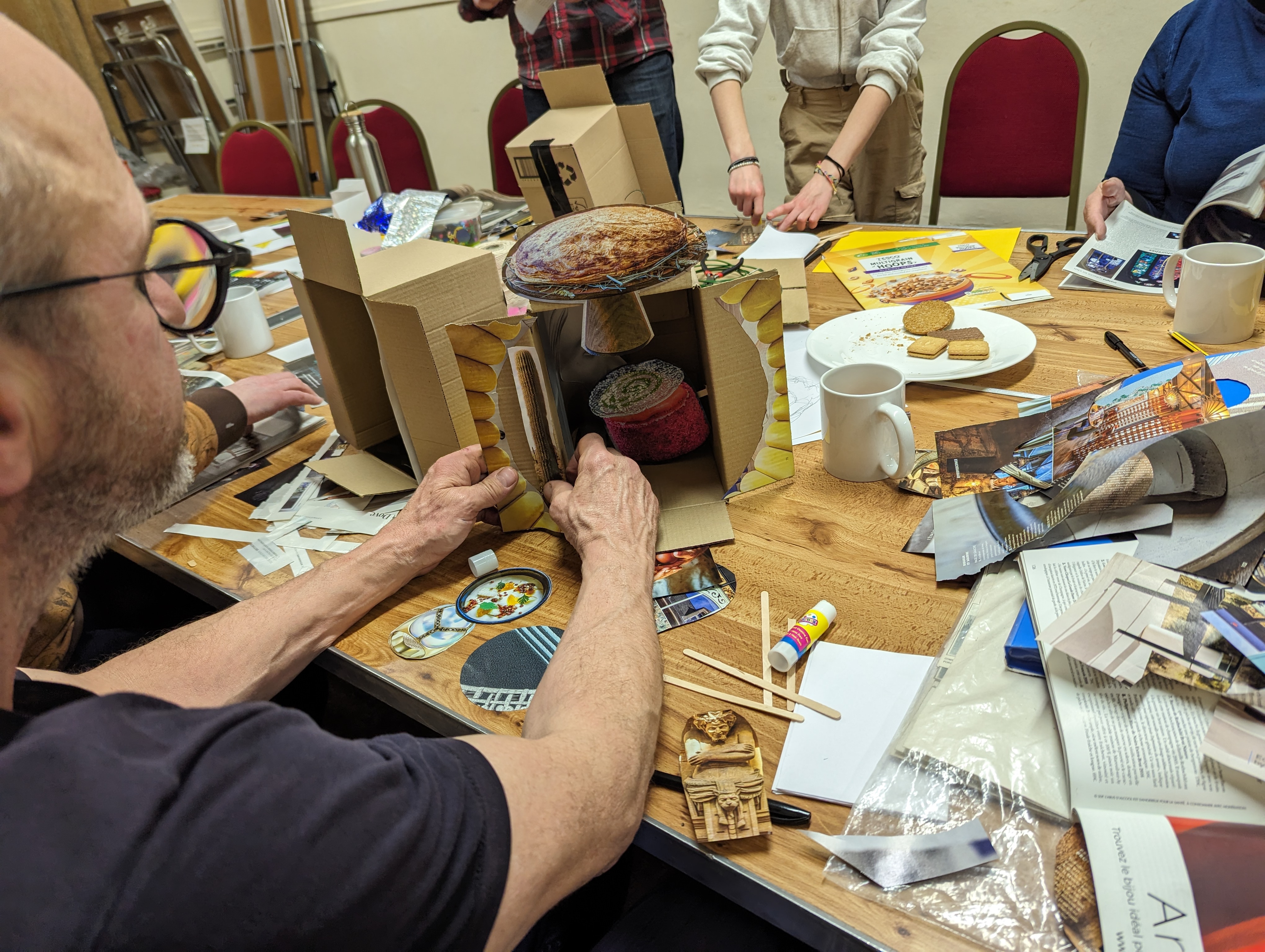 We see the shoulders of a man making a piece of artwork at a table. He is surrounded by magazine cuttings.