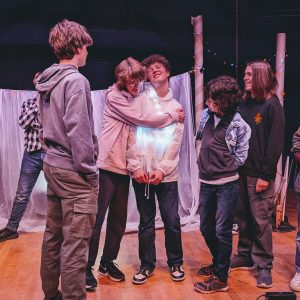 A group of 5 young people in a theatre production. Two boys are hugging and the other 3 people look on