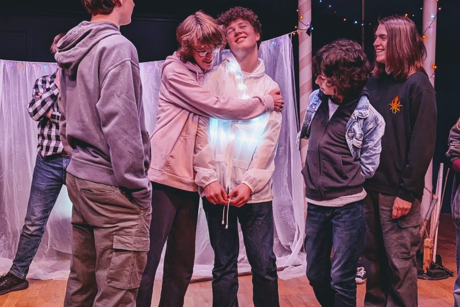 A group of 5 young people in a theatre production. Two boys are hugging and the other 3 people look on