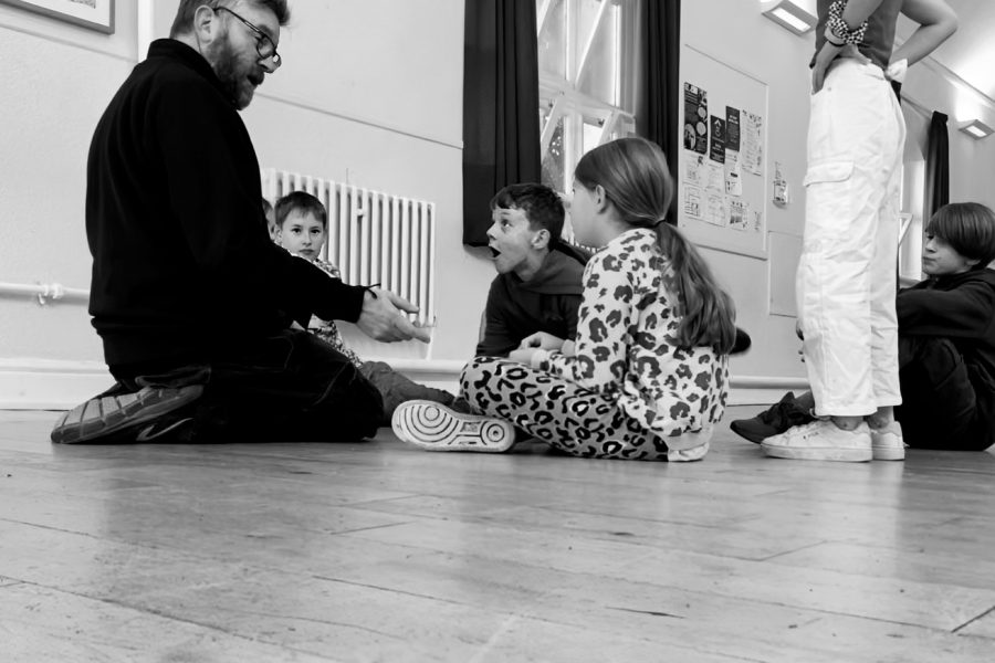 The image is black and white A workshop leader sits on a wooden floor surrounded by 5 children