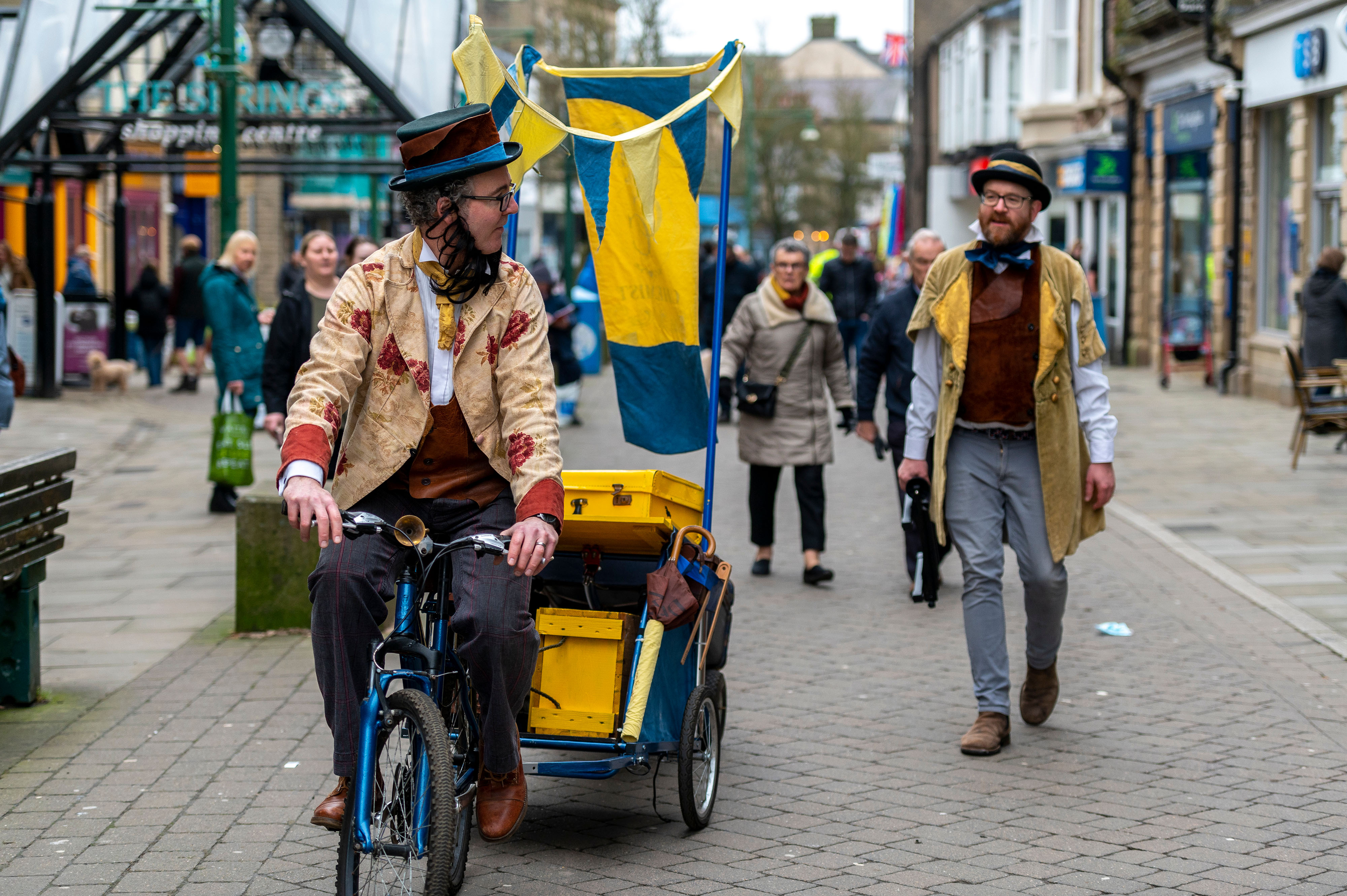 two men in victorian dress wallk a high street, one is riding a bike while the other walks alongside