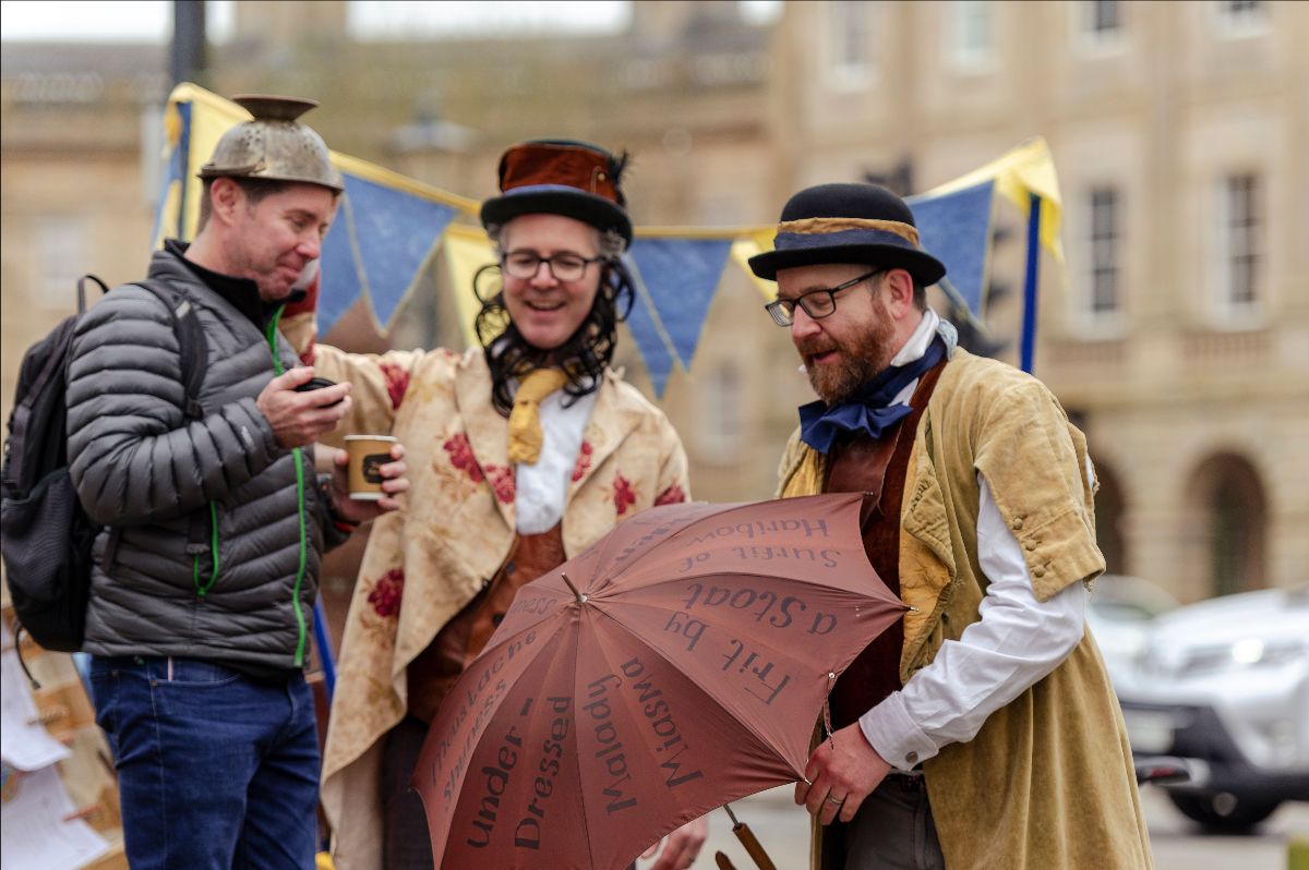 Two men dressed in Victorian style costume interact with a shoppper on a high street. They are all smiling