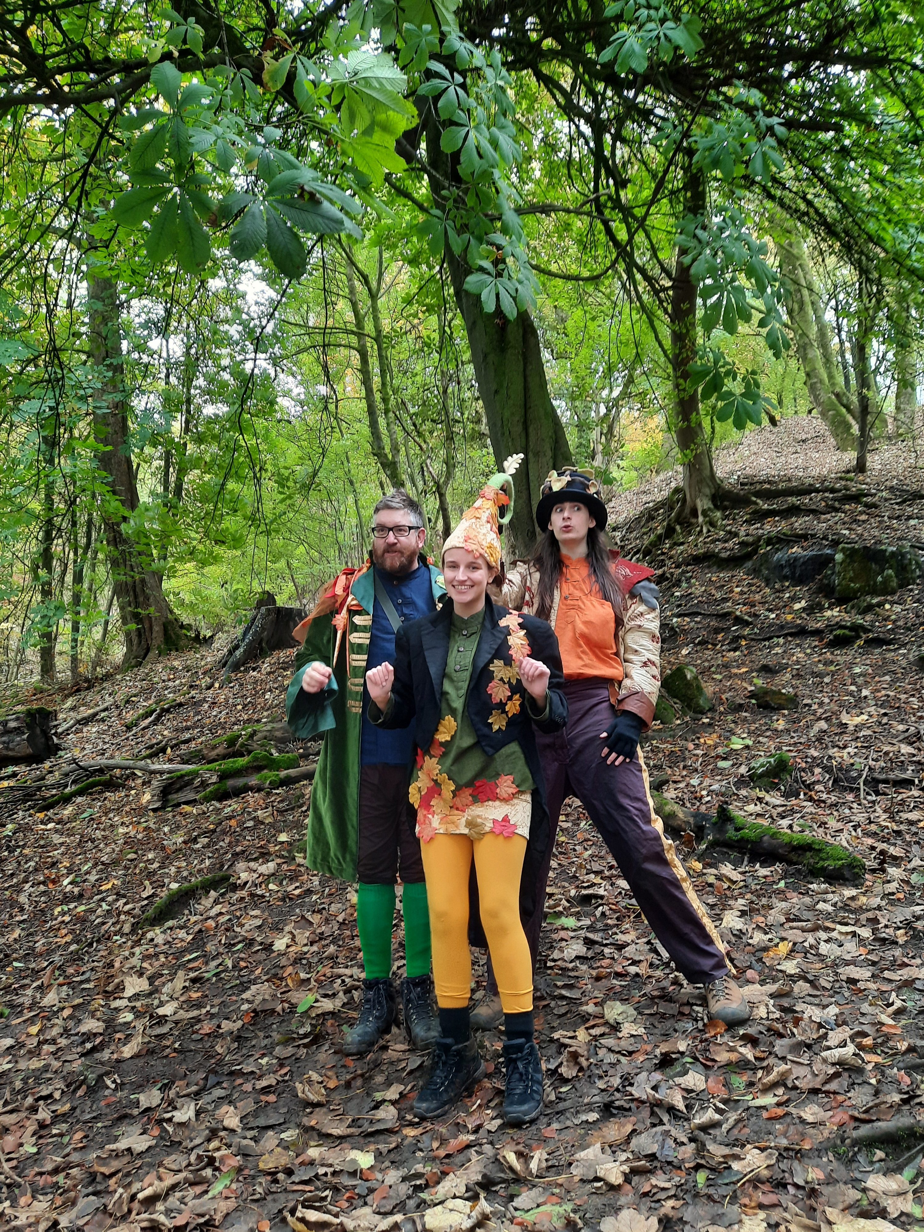 Three characters dressed in hats and clothing with a woodland theme stand together and smile at the camera