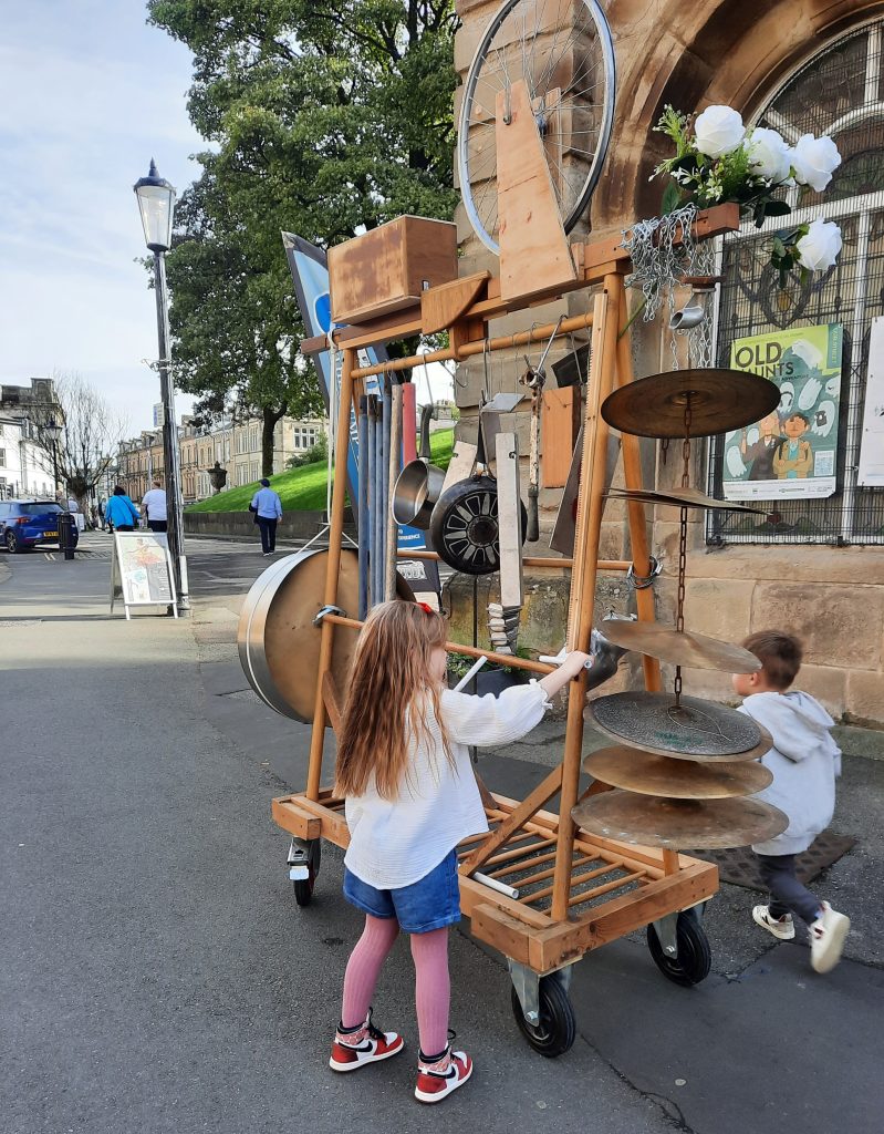 A young girl and boy play with a large junk style music set. It has pipes, pans and cymbals for everyone to play and bash