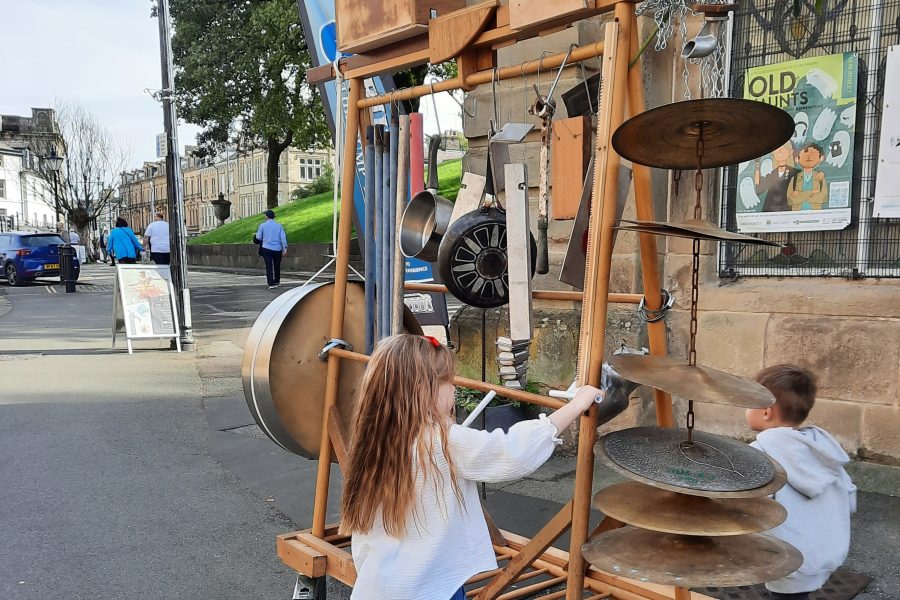 A young girl and boy play with a large junk style music set. It has pipes, pans and cymbals for everyone to play and bash