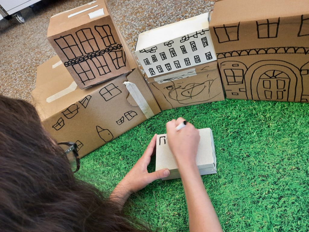 A child draws with a felt tip onto a small cardboard box to be part of a collection of small boxes that form a building