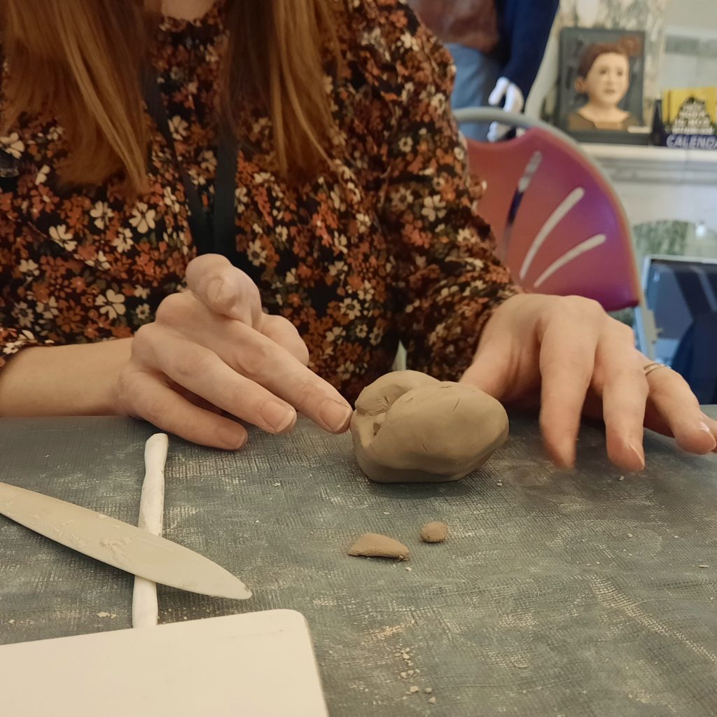 A woman sits at a table and sculpts some clay with her hands. There are clay tools on the table to her right.