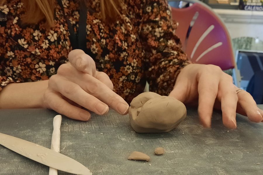 A woman sits at a table and sculpts some clay with her hands. There are clay tools on the table to her right.