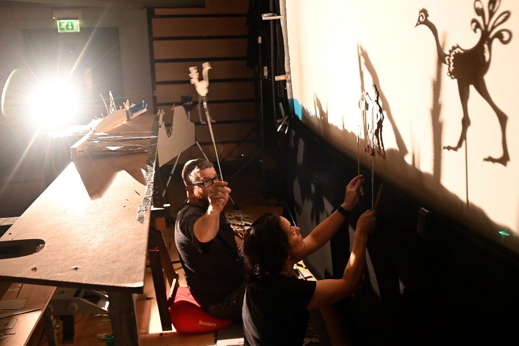 Two puppeteers are behnd a shadow screen holding up puppets. It s dark except for a source of light from the left illuminating the screen and puppets.