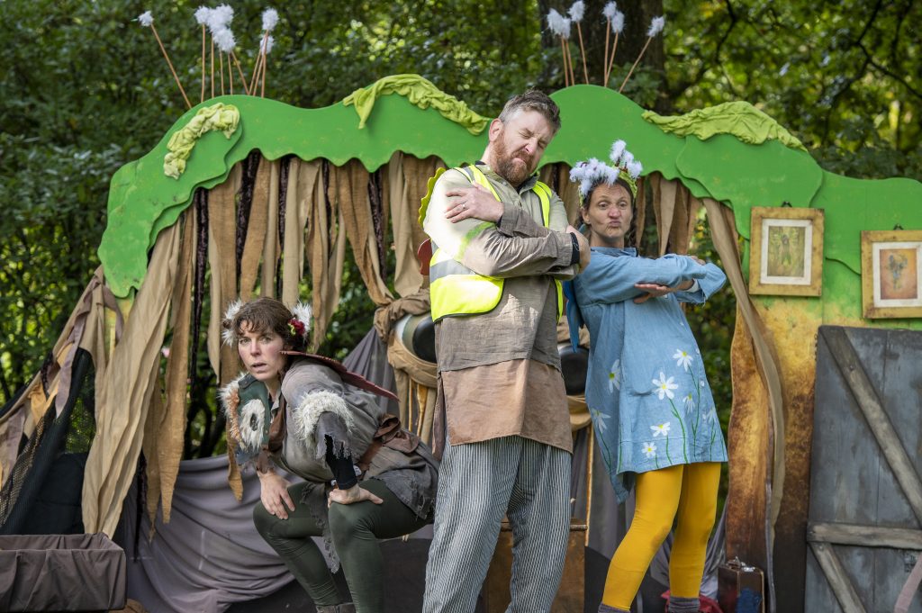 Three actors on an outdoor stage. They have attitude as they strike poses. Two are stood together arms folded, the other female actor crouches down.