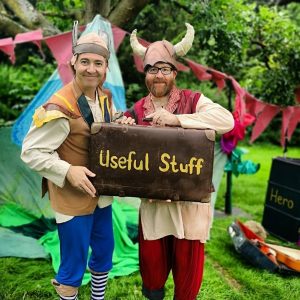 Two actors pretend to be monster menders. They are dressed up in hats, waistcoats and colourful trousers. They are holding an old fashioned leather suitcase that has the words Useful Stuff painted onto it.