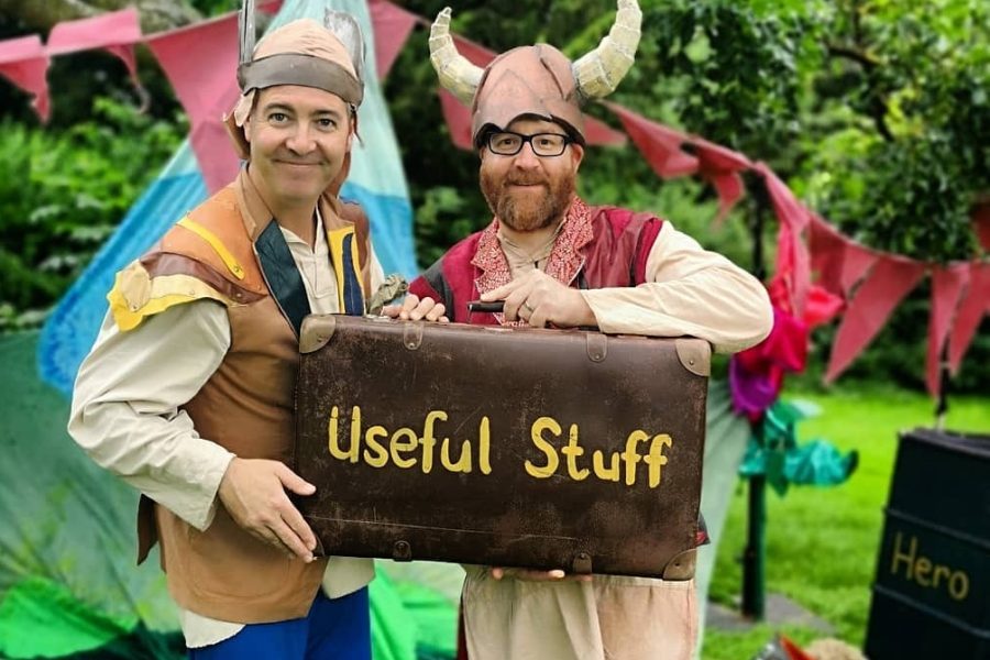 Two actors pretend to be monster menders. They are dressed up in hats, waistcoats and colourful trousers. They are holding an old fashioned leather suitcase that has the words Useful Stuff painted onto it.