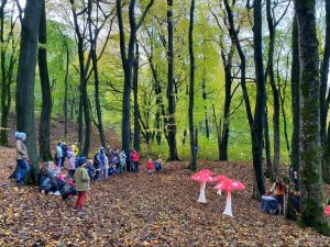 An audience gather in a woodland and there are brown leaves on the ground. They are watching a theatre show and there are big red mushrooms as the set.