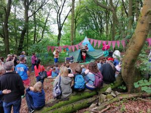 The photo is taken from behind the audience. They are sat in a woodland and are watching a theatre show about a dragon