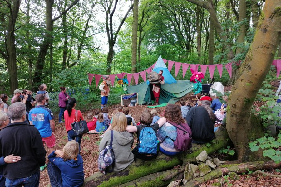 The photo is taken from behind the audience. They are sat in a woodland and are watching a theatre show about a dragon