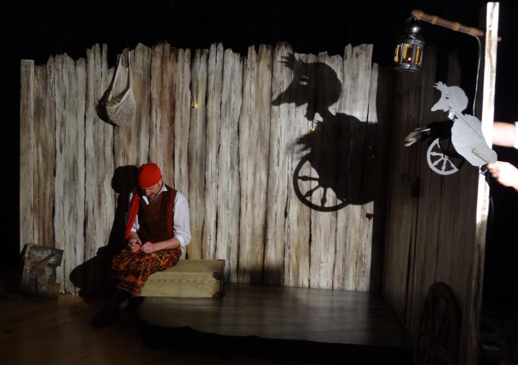 An actor sits on a semi dark stage in a red hat, next to a suitcase. There is a shadow on the wall behind them of rumpplestiltskin