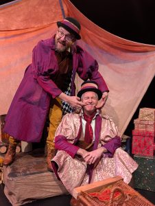 Two actors are wearing pink and purple flambouant frock coats. One sits on the edge of the stage and the other is stood behind them trying to make their mouth lift into a smile