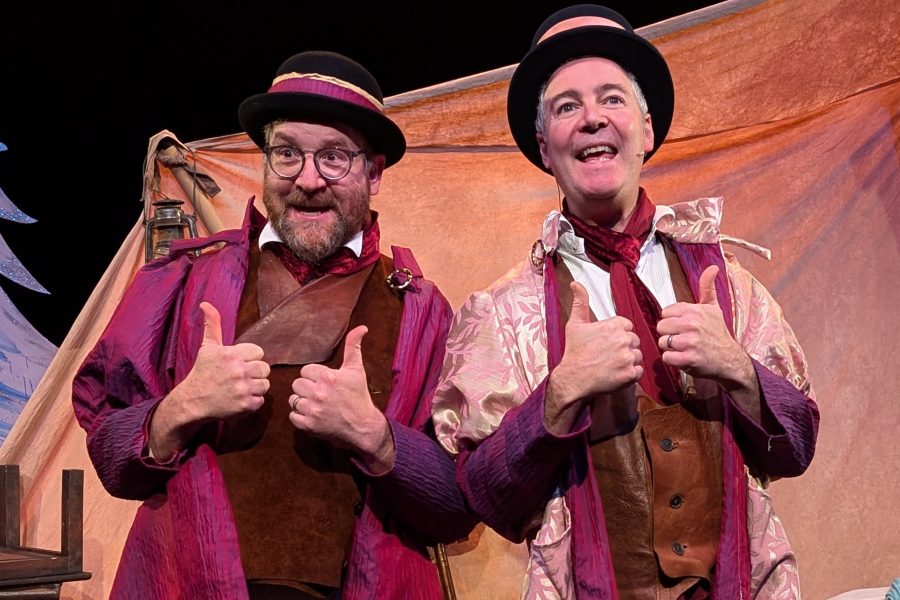 Two actors pretending to be brothers are dressed in fine clothes and pink and purple flamboyant jackets. They are looking out and smiling with their thumbs up.