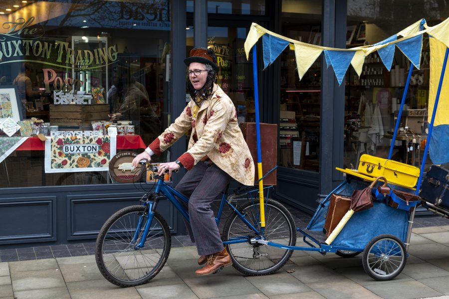 a man dressed in a colourful floral coat rides a bicycle pulling a small trailer that has poles and bunting