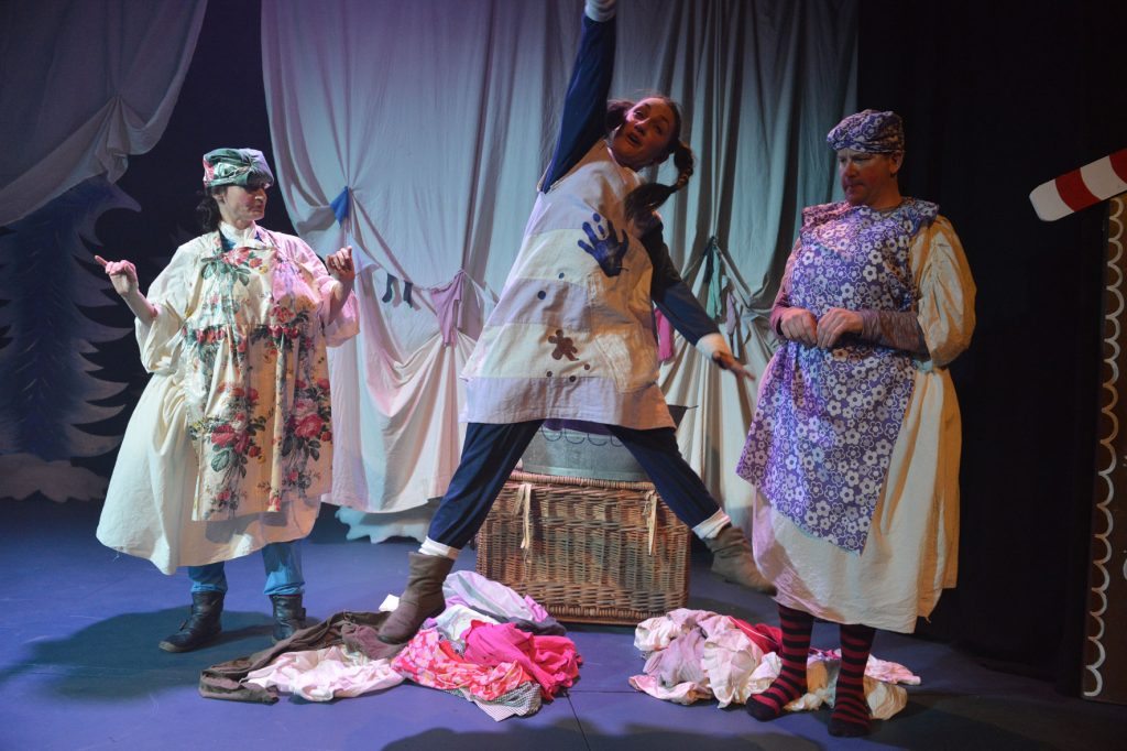 Three actors on stage amongst a pile of laundry. Two washer women and a young girl. The young girl is leaping in the air fist held high