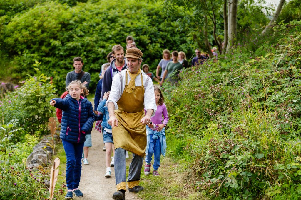 A male actor in a white shrt and gold apron and hat leads a family audience along a pathway through green bushes