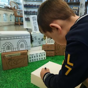 A young child draws on a small cardboard box. It's part of a craft activity where particpants are making a buildings of a town.