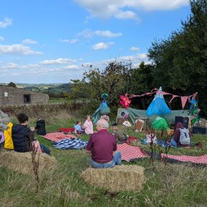 A theatre set has been set up in a farmers field and families are sat on hay bales. The sky is blue and there are white fluffy clouds.