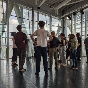 A group of young people stand together getting ready for a theatre workshop. They are in a bright room with glass windows.