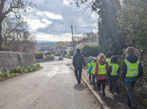 School children walk down a street in their village. They are wearing illuminos jackets. An adult is leading them down the road and the sun is shining.