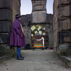 A man dressed in a three quarter length purple dress coat with bowler hat stands at the entrance of a stone walled building. Through the entranceway we can see an elabourate dresser illuminated in the dusky evening